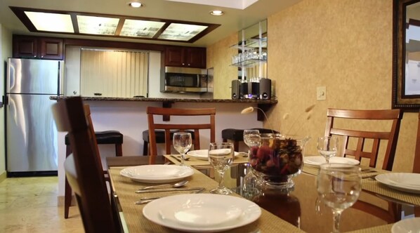 Un comedor con una mesa dispuesta para cuatro, con platos, cubiertos y copas de vino. Se observan un microondas y un refrigerador al fondo.