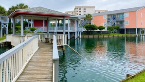 A waterfront property with a dock leading to a covered area, adjacent buildings, and a calm water body. - 🌟 New 🌟 Fun Mid-Century Modern Beachside Condo🏝🐠 (Pensacola Beach)