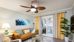 A living room with a brown sofa, a glass coffee table, a ceiling fan, a large window with yellow patterned curtains, and a potted plant. - 🌟 New 🌟 Fun Mid-Century Modern Beachside Condo🏝🐠 (Pensacola Beach)