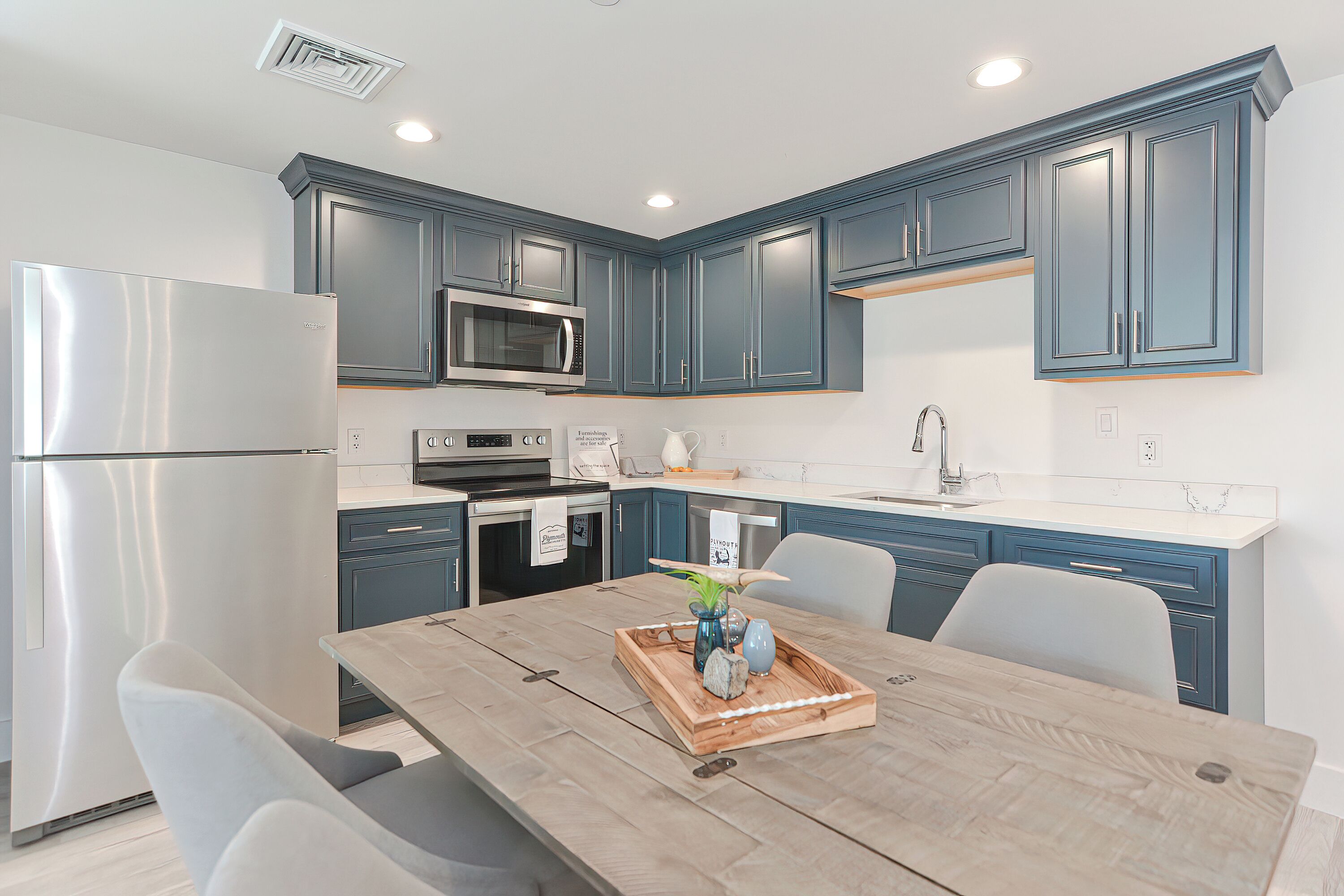 A modern kitchen with blue cabinets, stainless steel appliances, and a wooden dining table with a tray of decorative items.