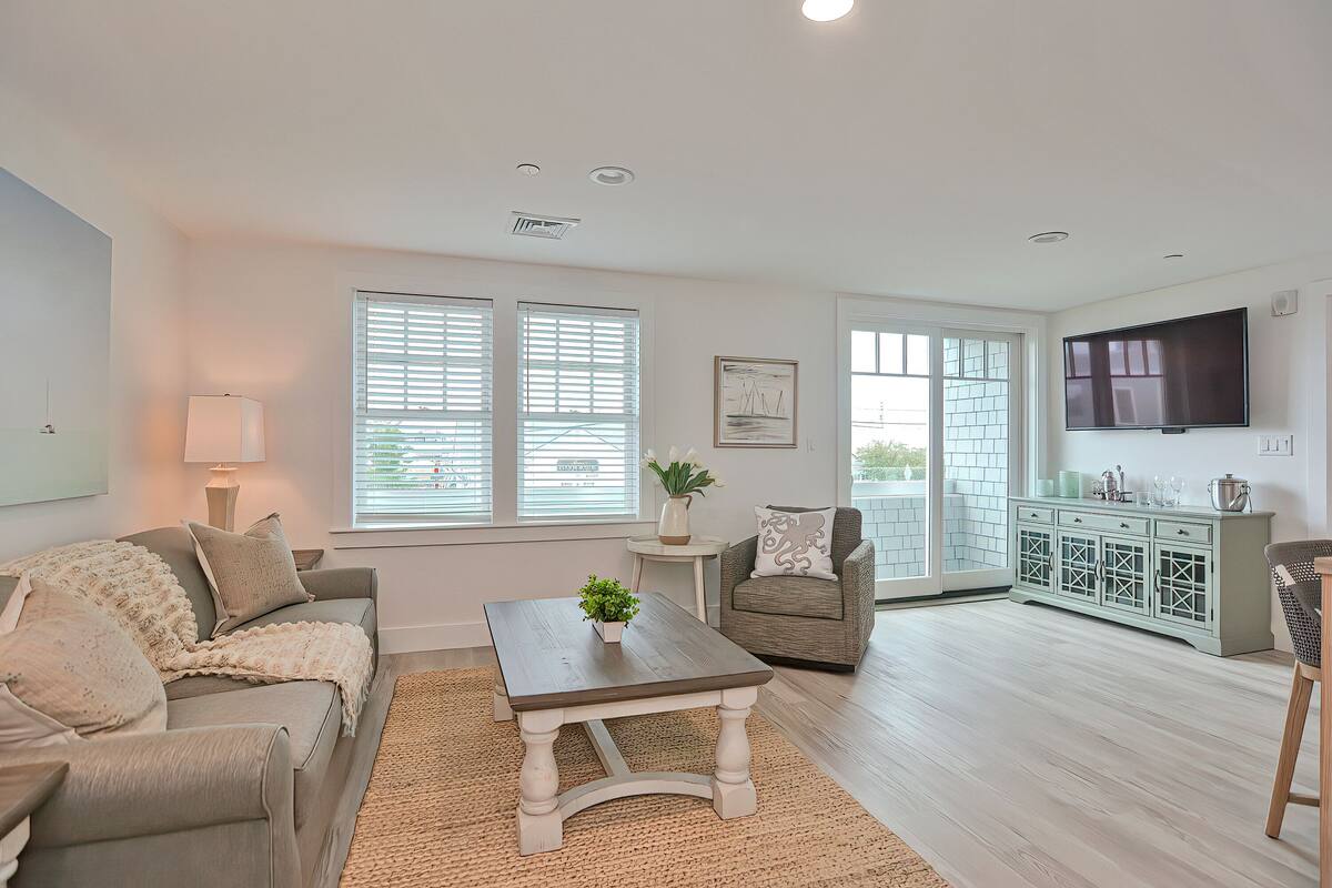 A living room with a sofa, coffee table, and a television. There are windows with blinds, a small table with a plant, and a cabinet with decorative items.