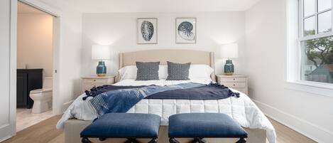 A bedroom with a bed, two bedside tables, a window, and a door leading to the bathroom.