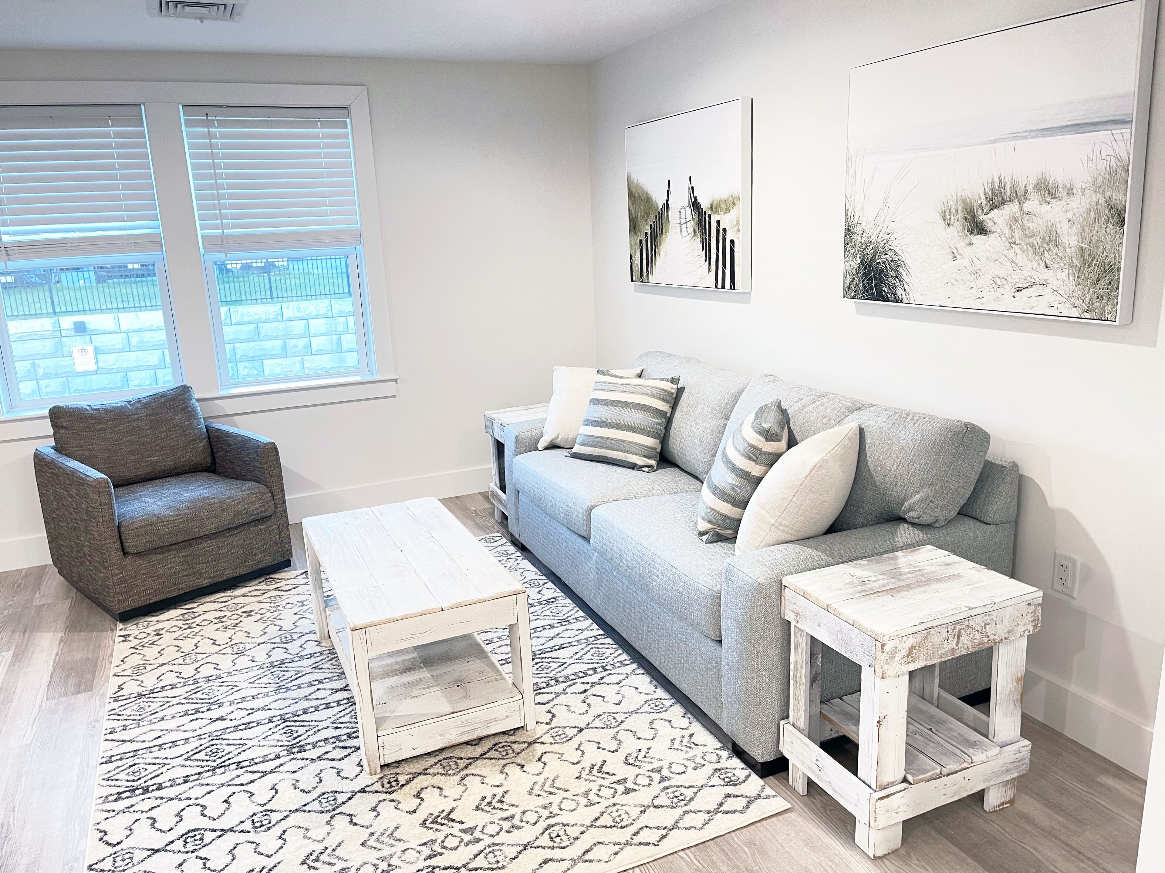Una sala de estar con un sofá gris, un sillón gris, una mesa de centro blanca y una mesita auxiliar blanca. Hay dos ventanas con persianas, una alfombra estampada y dos cuadros colgados en la pared.