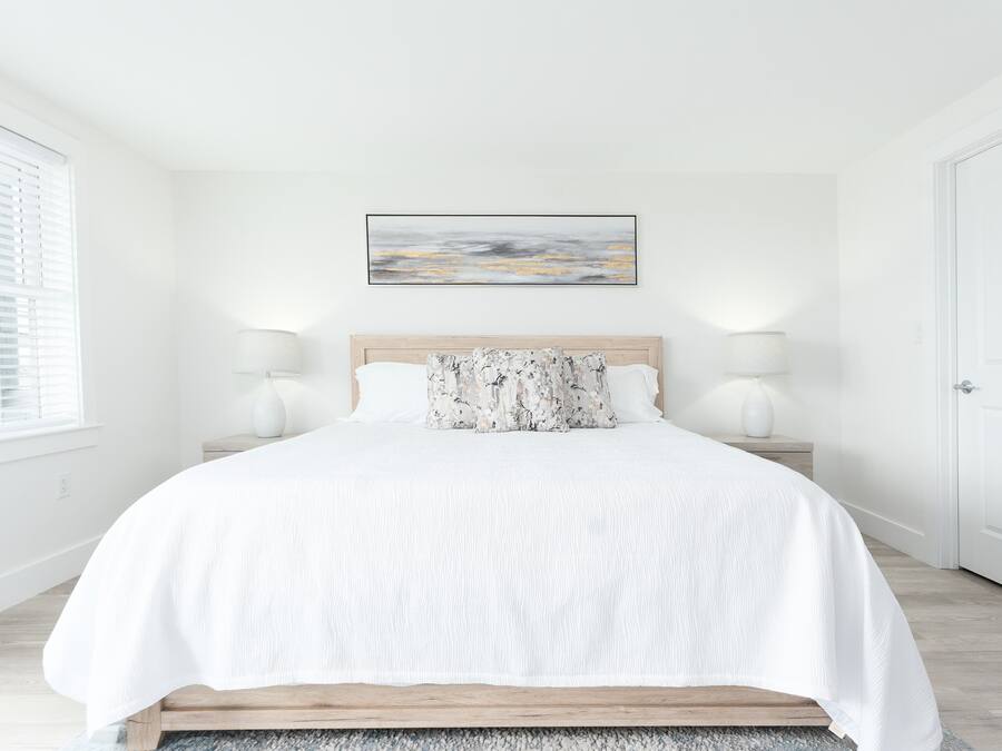 Een slaapkamer met een bed, witte beddengoed, twee bedlampen, een houten hoofdbord, een schilderij aan de muur, en een raam met jaloezieën.