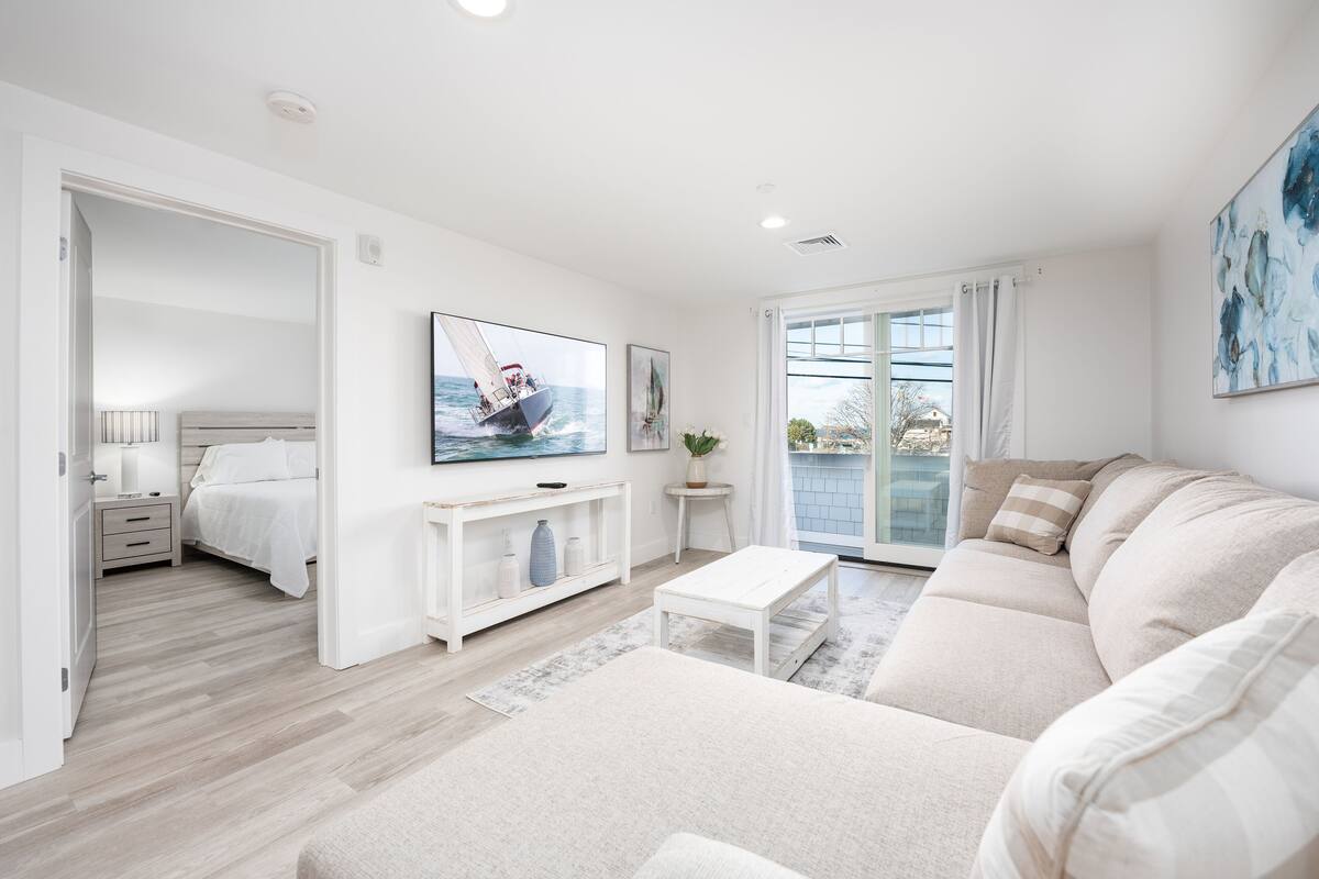 A modern living room with a sofa, a coffee table, and a TV. There is a bed in the adjacent room and a balcony with a view.