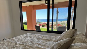 A bed with a patterned bedspread and pillows, a window with a view of a garden and the ocean, and a ceiling fan. - Beach Front Pool Fully Kitchen Breathtaking Sea Views Close to Everything (San Jose Del Cabo)