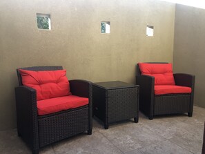 Two wicker chairs with red cushions and a small table, set against a plain wall with two small windows.