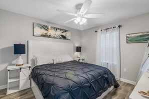 Une chambre à coucher avec un lit, un ventilateur de plafond, une table de chevet avec une lampe et une fenêtre avec des rideaux.