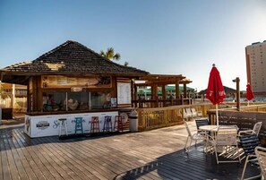 Un petit kiosque situé sur la plage, doté d’un toit en bois, d’un panneau affichant le menu et d’un parasol à l’effigie de Coca-Cola.