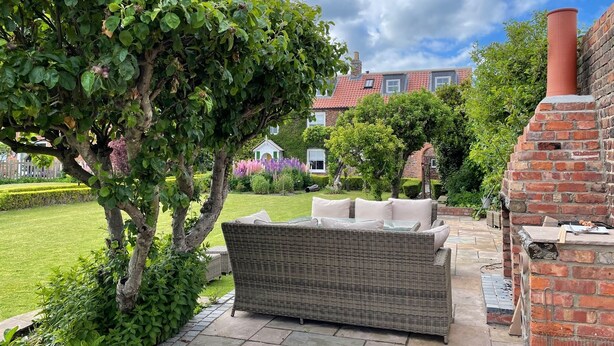 A garden patio with a wicker sofa, a brick barbecue, and a lawn with a hedge and trees.