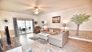 A spacious living room with a ceiling fan, a large window with curtains, a sofa, a coffee table, and a television. - Lake & Pool View in the Heart of Myrtle Beach, SC-Magnolia Pointe-Myrtlewood (Myrtle Beach)