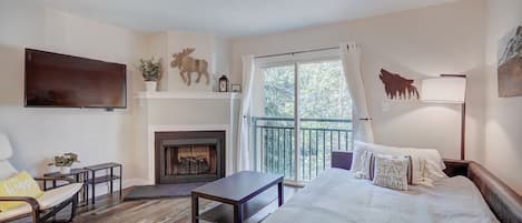 A cozy room with a fireplace, a sofa, a TV, and a balcony with a view of trees.