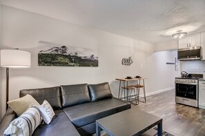 Relax in this stylish living area with a sofa, ottoman, and dining table, surrounded by a large landscape painting and sleek kitchen.