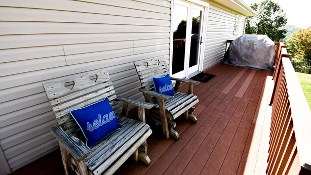 Una terraza de madera con dos sillas Adirondack, cada una con un cojín azul, y una parrilla cubierta a la derecha.