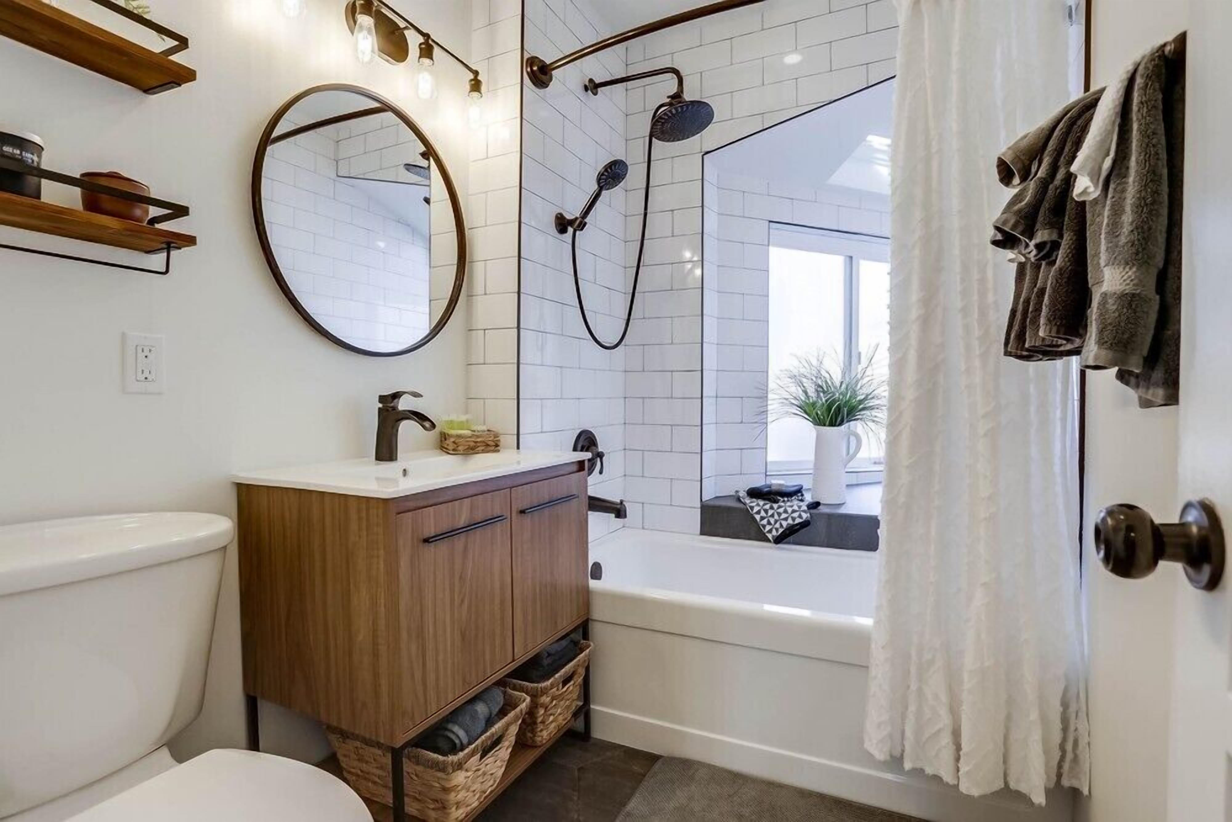 A modern bathroom with a white toilet, wooden vanity, round mirror, and a bathtub with a showerhead.