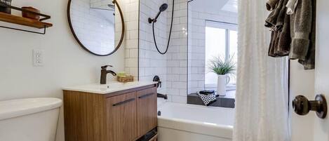 A modern bathroom with a white toilet, wooden vanity, round mirror, and a bathtub with a showerhead.