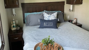 Une chambre à coucher comprenant un lit, une table de chevet, une lampe, une étagère murale décorée de divers objets, et un panneau décoratif au-dessus du lit.