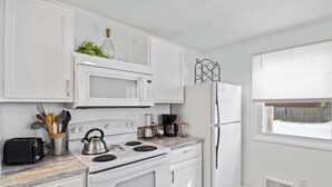 A modern kitchen with white cabinetry, a stove, microwave, and refrigerator. There is a window with blinds, a toaster, and a kettle on the stove.