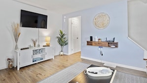 A modern living room with a flat-screen TV, a white console table, a coffee table, and a wall-mounted shelf with a laptop and a lamp.