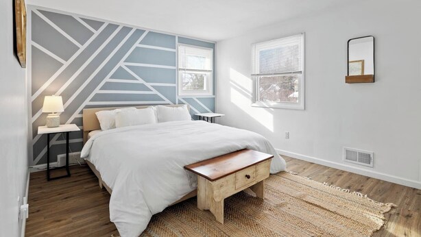 A bedroom with a bed, a nightstand, a lamp, a window with blinds, a mirror, and a geometric wall design.