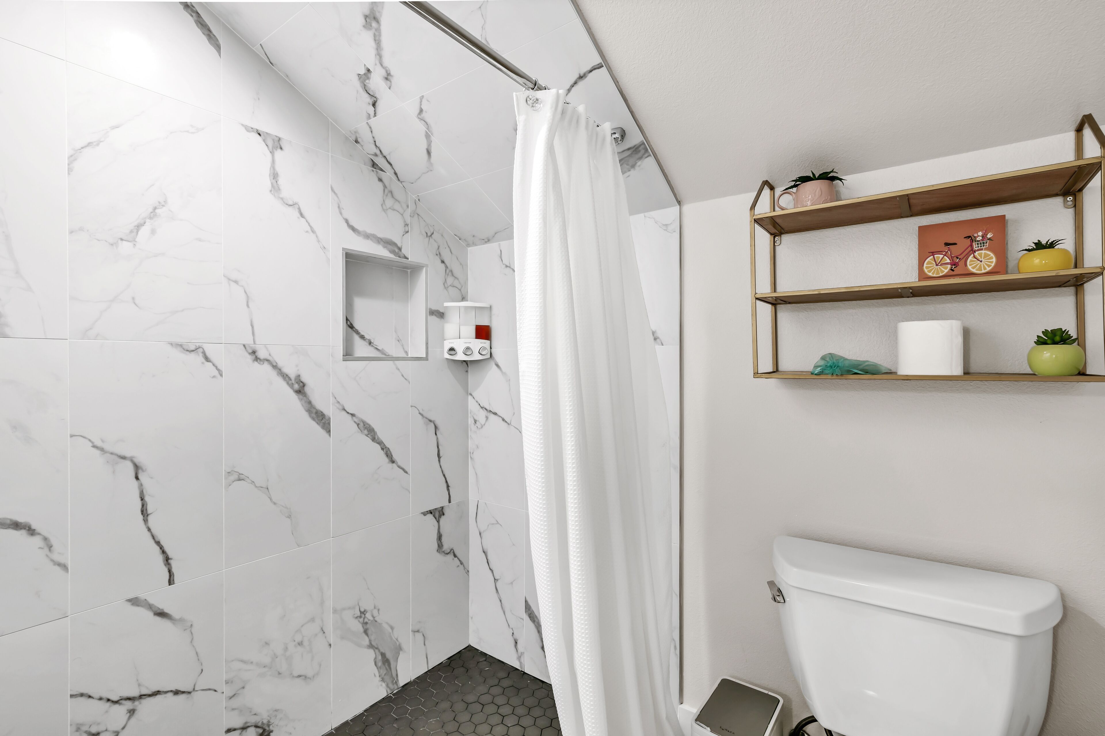Un baño moderno con azulejos de mármol blanco, ducha con cortina blanca, inodoro y un estante con artículos de aseo.