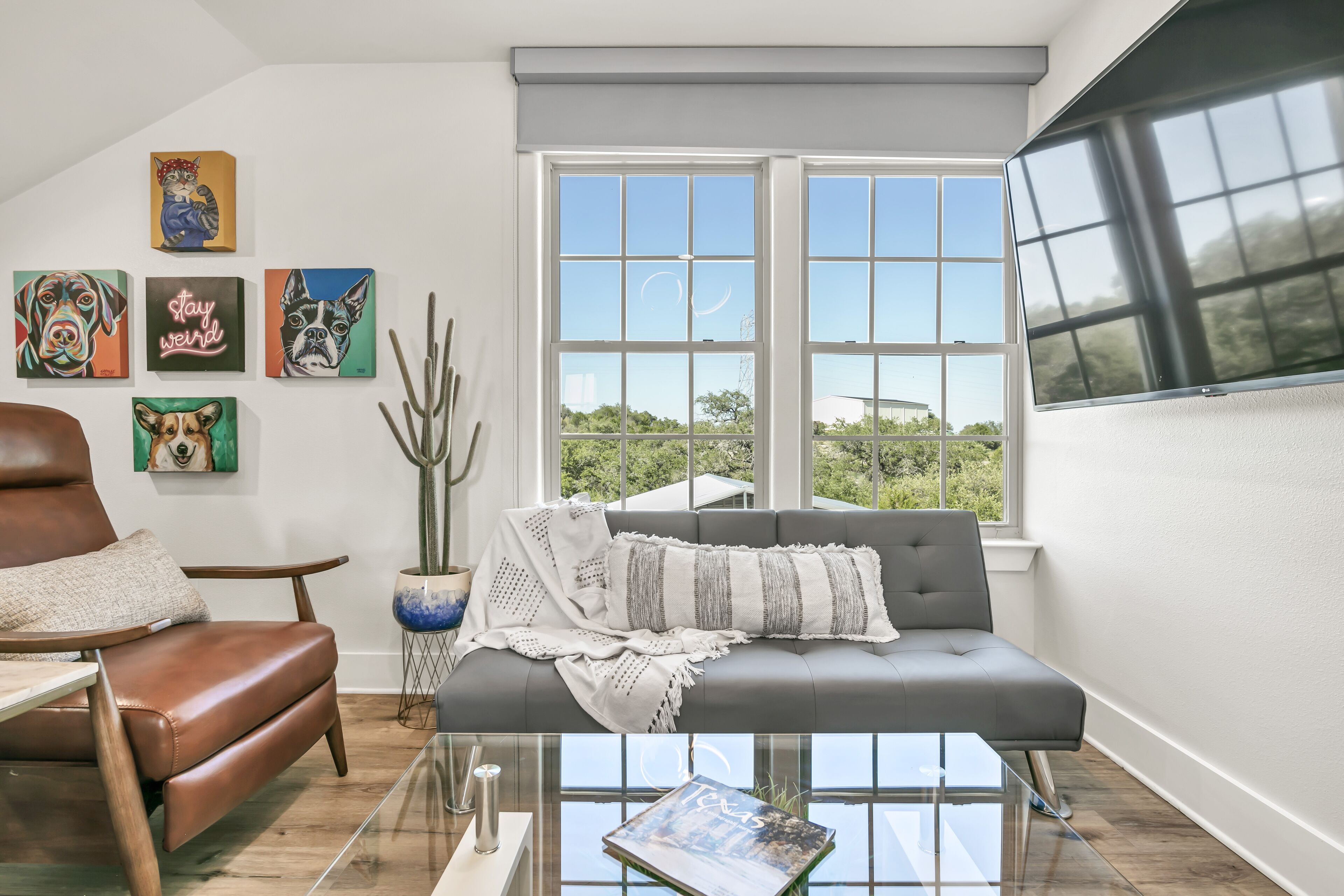 A modern living room with a grey sofa, a brown leather chair, and a glass coffee table. There are six framed artworks on the wall, and a large window with a view of greenery outside.