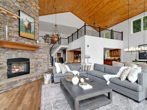 A spacious living room with a stone fireplace, a grey sectional sofa, a dark coffee table, and a staircase leading to an upper level.