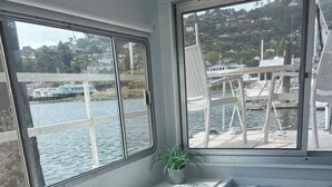 A room with a bed, a window with a view of a body of water and buildings, a small table with a tissue box and a green cup, and a potted plant.