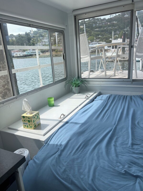 A room with a bed, a window with a view of a body of water and buildings, a small table with a tissue box and a green cup, and a potted plant.