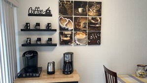 Een compacte kitchenette met een koffiezetapparaat, waterkoker en een koffiefilter op het aanrecht. Daarboven staan planken met mokken en een wanddecoratie met koffiethema.