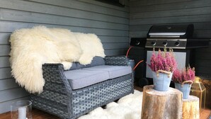 Un'accogliente area esterna con un divano in rattan grigio, una soffice coperta bianca e un tappeto morbido bianco. Due fioriere in metallo con fiori rosa su sgabelli di legno. Sul pavimento, un portacandele in vetro con pietre bianche.