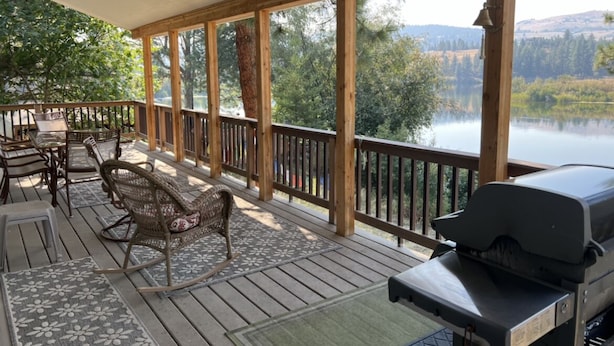 A covered deck with outdoor furniture, a grill, and a view of a lake and forest. - Charming Waterfront Pine Cabin on Curlew Lake (Republic)