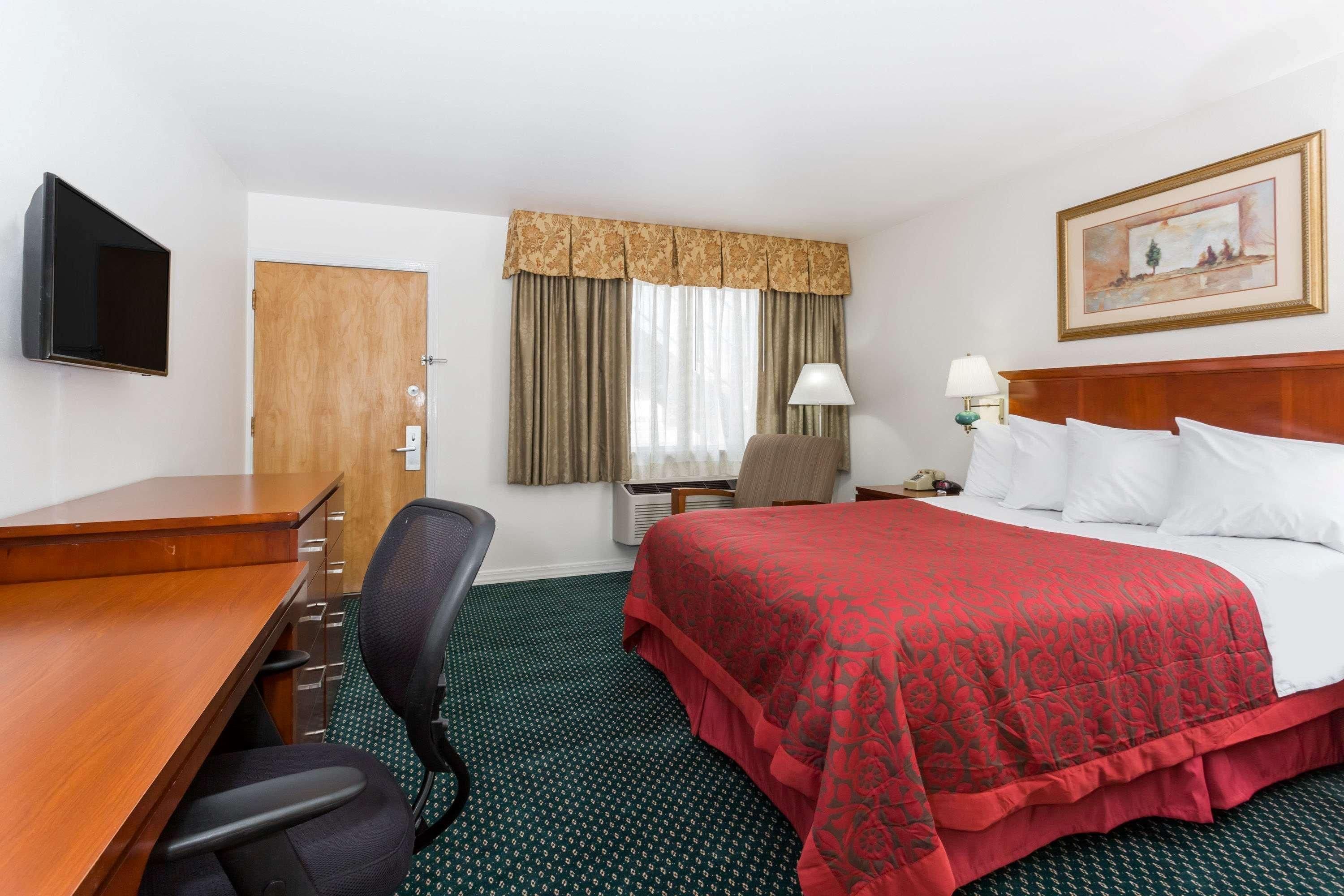 Une chambre d’hôtel avec un bureau en bois, une chaise, un lit recouvert d’une courtepointe rouge, une table de chevet avec une lampe, un tableau au mur et une fenêtre avec des rideaux.