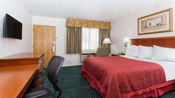 Une chambre d’hôtel avec un bureau en bois, une chaise, un lit recouvert d’une courtepointe rouge, une table de chevet avec une lampe, un tableau au mur et une fenêtre avec des rideaux.