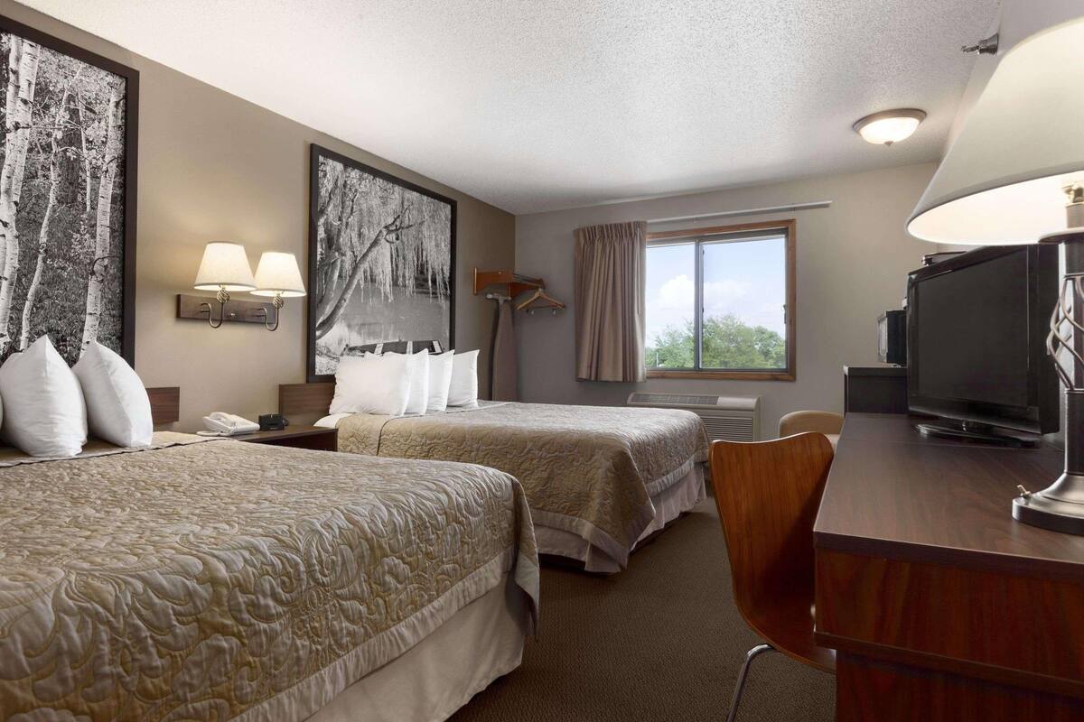A hotel room with two beds, a desk with a TV, a lamp, and a large window.