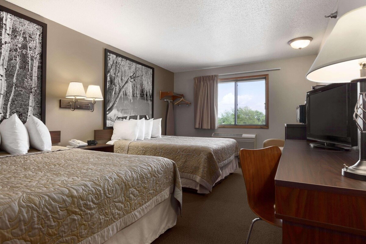 A hotel room with two beds, a desk with a television, a window with curtains, and artwork on the walls.