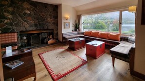 Een gezellige woonkamer met een stenen open haard, een bank met oranje kussens, een bijzettafeltje en uitzicht op een meer door grote ramen.