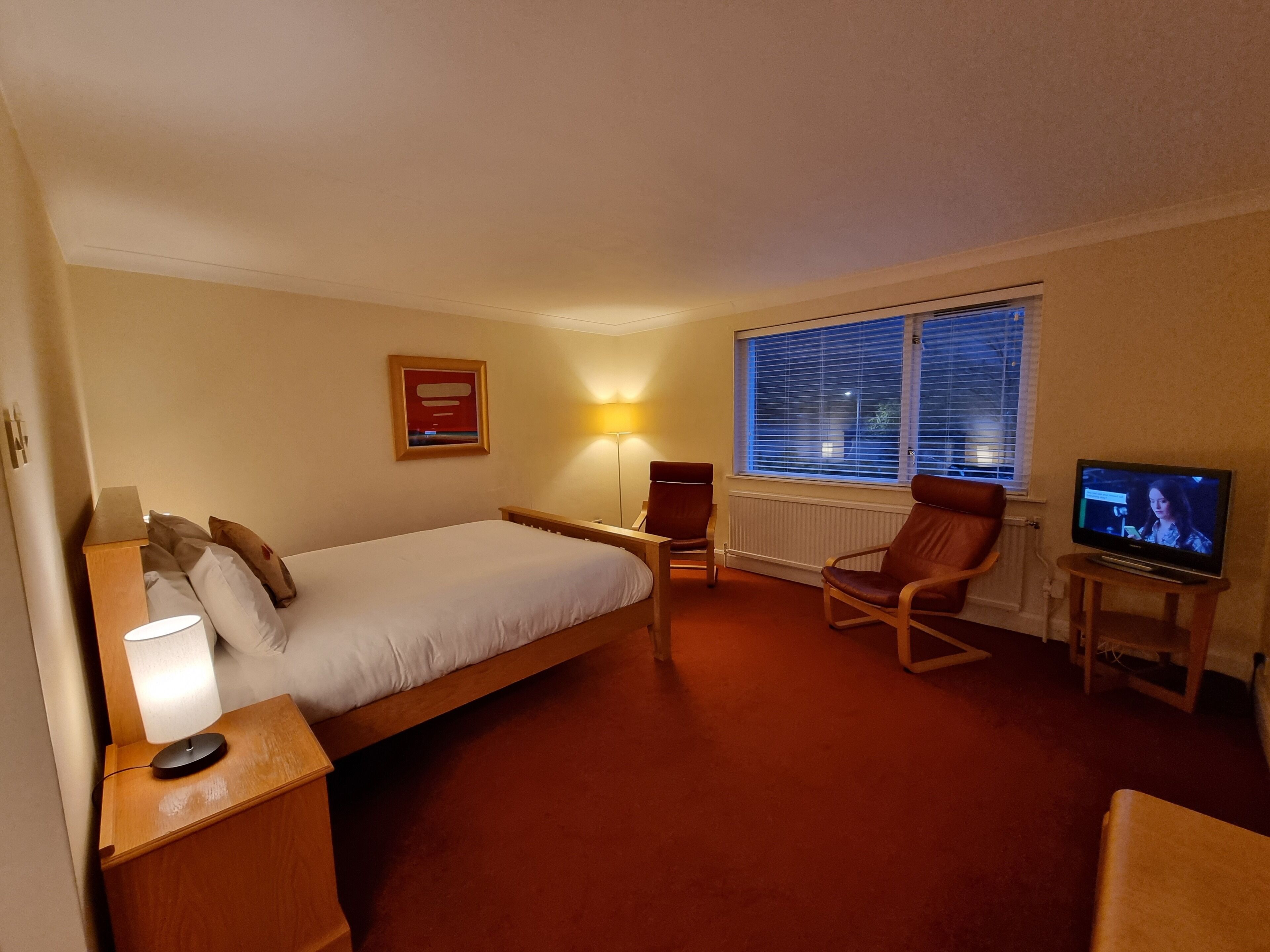 Een hotelkamer met een bed, een bureau met lamp, een stoel, een televisie en een raam met zonwering.
