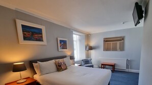 A hotel room with a bed, bedside table, lamp, chair, and artwork on the walls.
