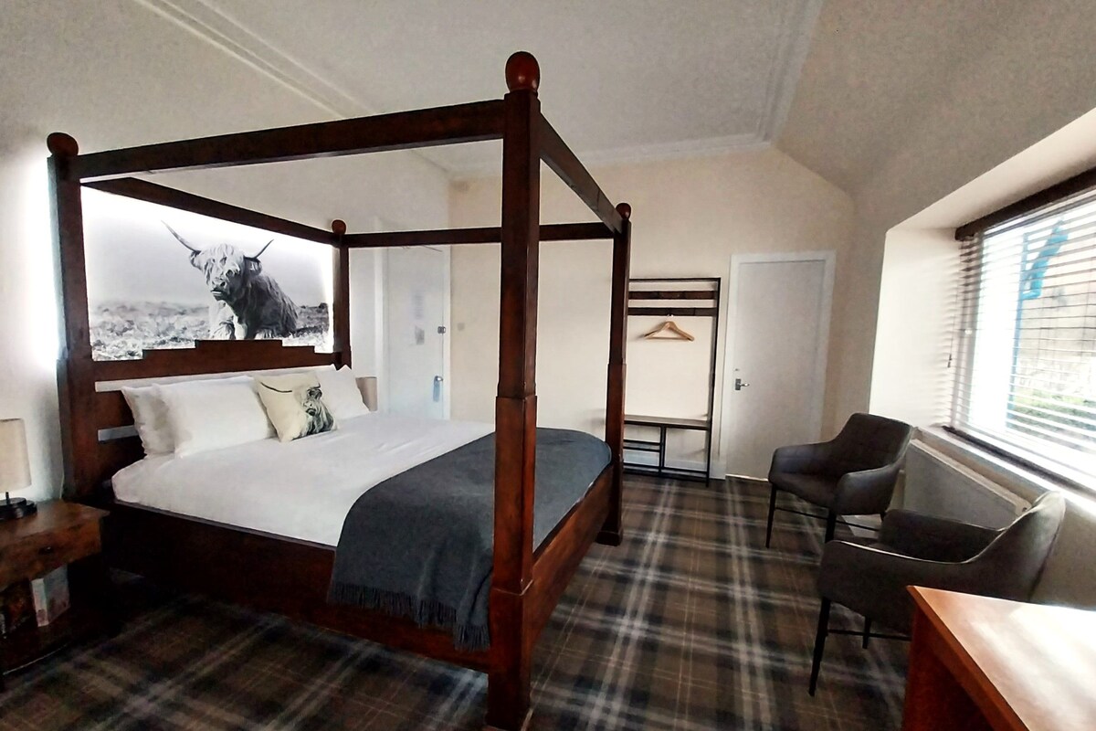A four-poster bed with a plaid carpet, a desk with a chair, and a coat rack in a hotel room.