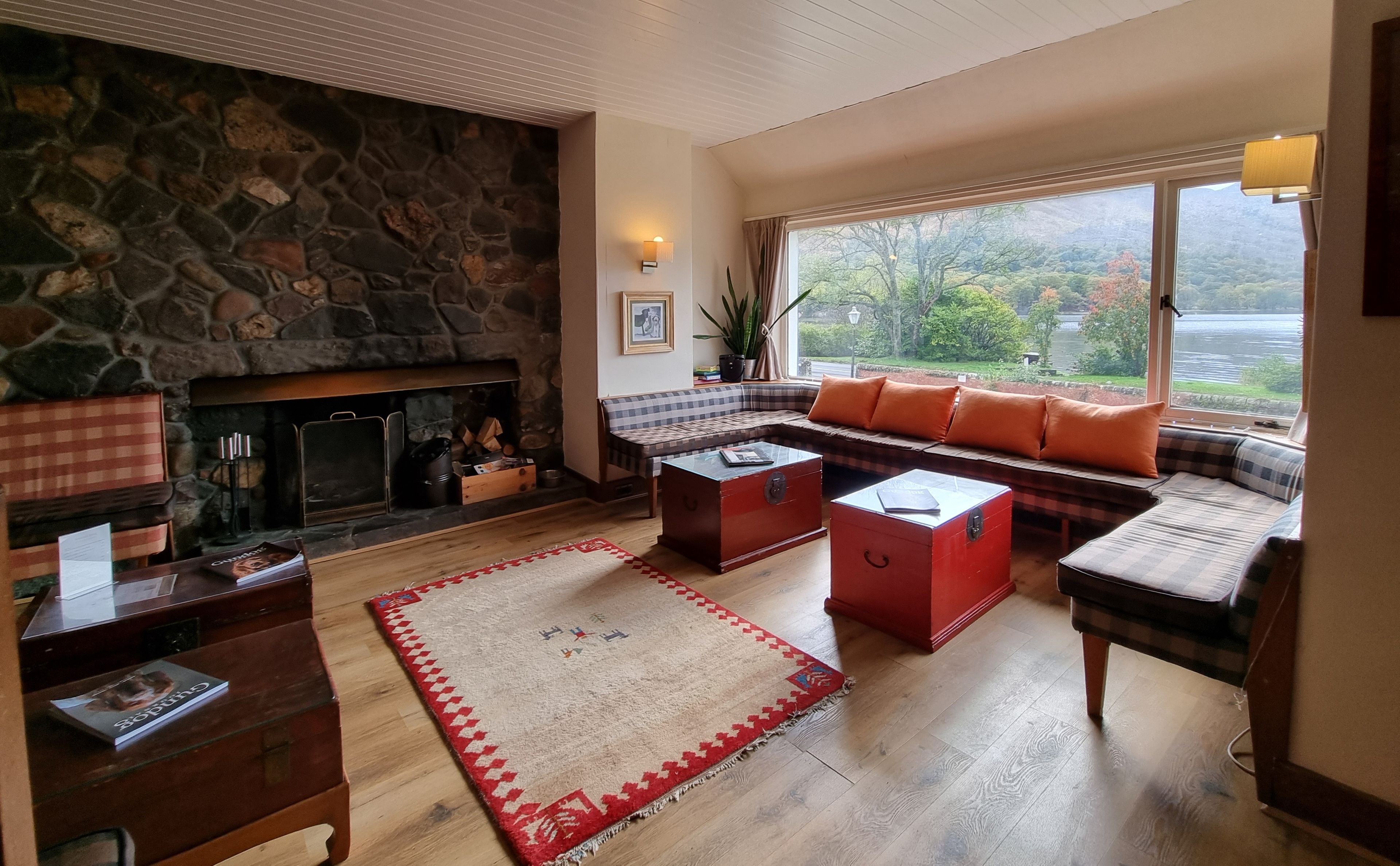 A cozy living room with a stone fireplace, a sofa with orange cushions, a coffee table, and a view of a lake through large windows.