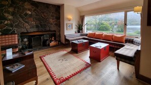 A cozy living room with a stone fireplace, a sofa with orange cushions, a coffee table, and a view of a lake through large windows.