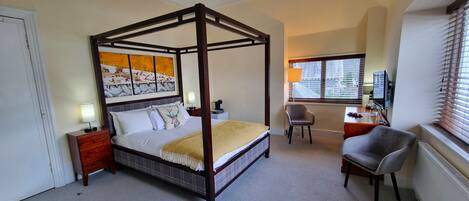 A four-poster bed with a headboard, a desk with a chair, a television, and a window with blinds.
