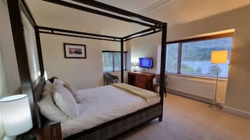 A four-poster bed with a headboard, a bedside table with a lamp, a desk with a computer, and a chair.