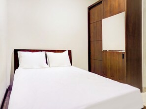Una cama con cabecera de madera y dos almohadas, un armario de madera con espejo, y una pared lisa.