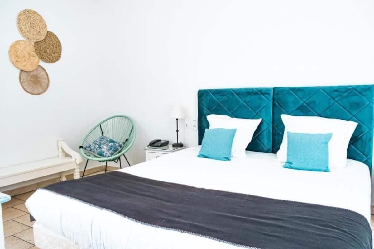Una cama con cabecera acolchada, una mesita de noche con lámpara, una silla y un adorno mural tejido.