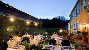 An outdoor dining area with tables set for guests, surrounded by greenery and a building with a balcony.