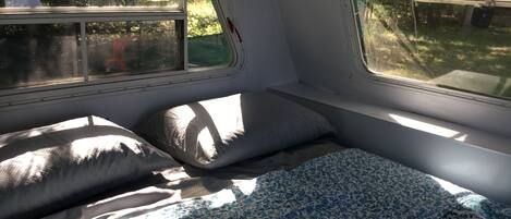 A compact RV interior with a bed, two windows, and a small bench.