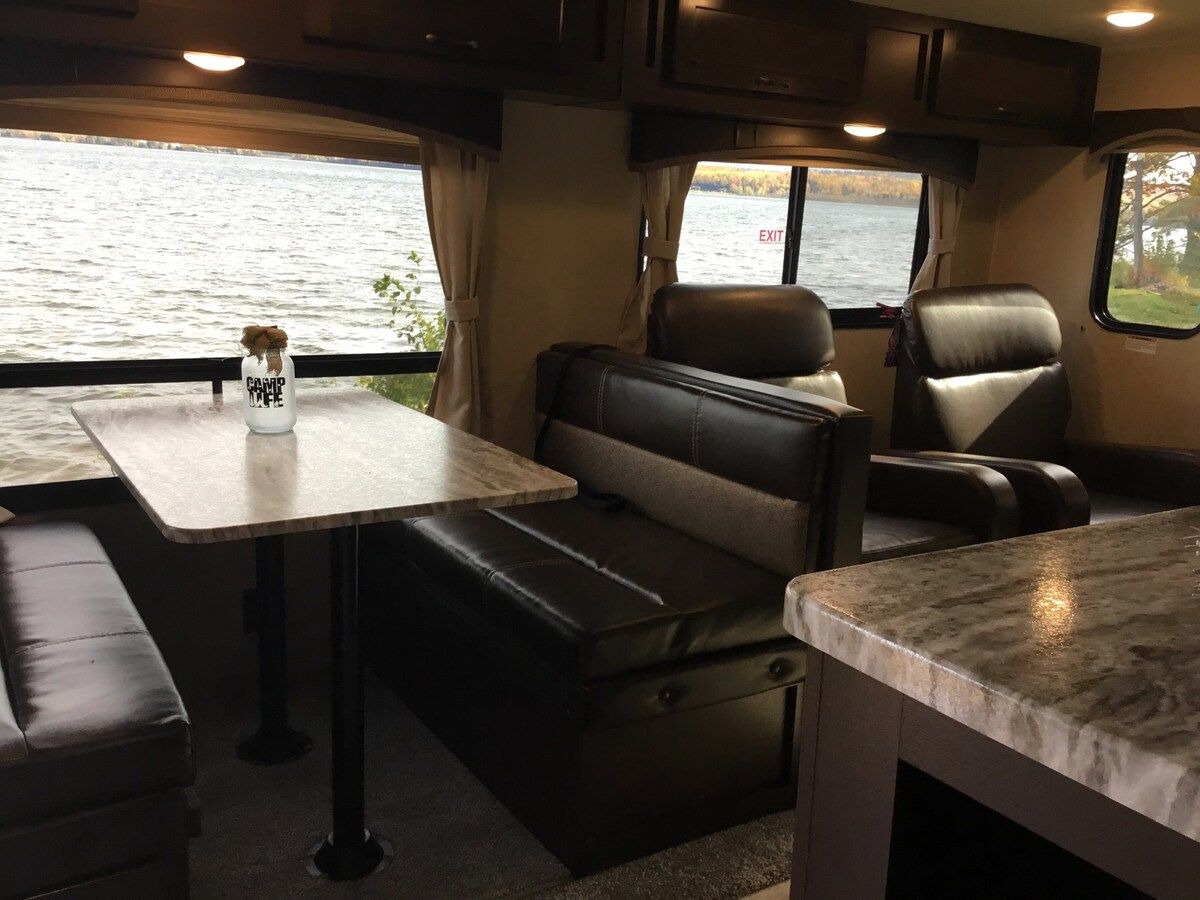 A compact RV interior with a dining area, seating, and a view of a body of water.
