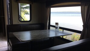 Een klein, afgesloten zitgedeelte met een tafel en bankjes, een raam met uitzicht op een wateroppervlak, en een fles met een etiket op de tafel.